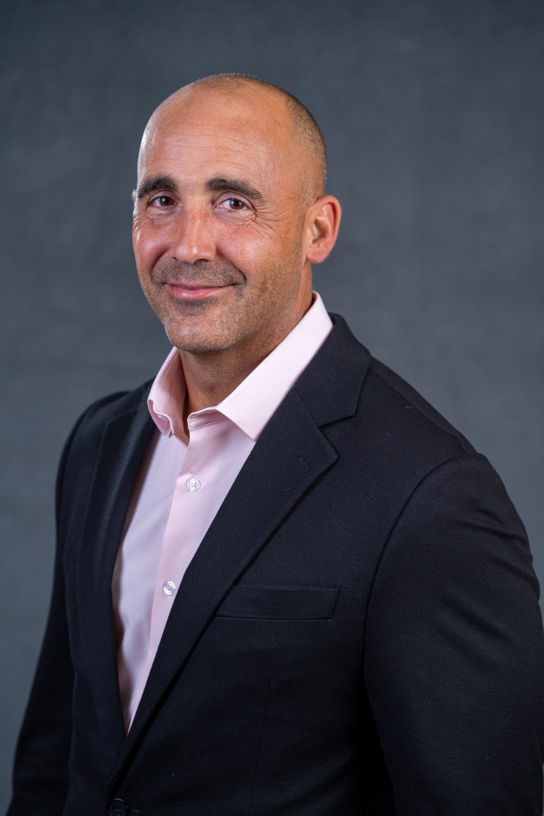 Headshot of a man in a dark suit and pink shirt against a dark gray backdrop