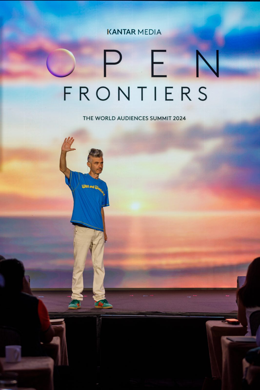 Speaker waving to the audience on stage under a giant sunset LED screen with the event logo
